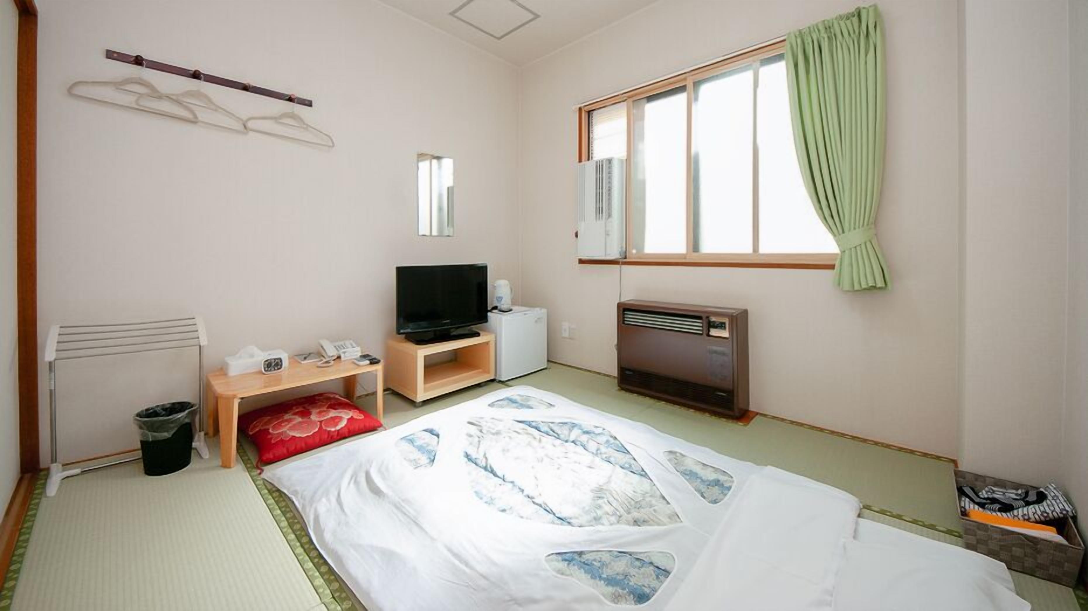 A hotel room with a bed, a TV, a heater, a radiator, a window with curtains, and a wall-mounted shelf.