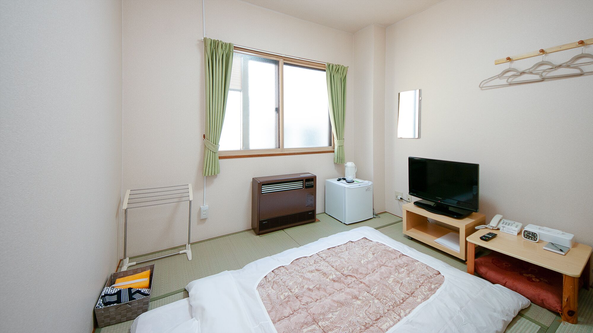 A hotel room with a bed, a television, a heater, a small table with a phone, and a window with curtains.
