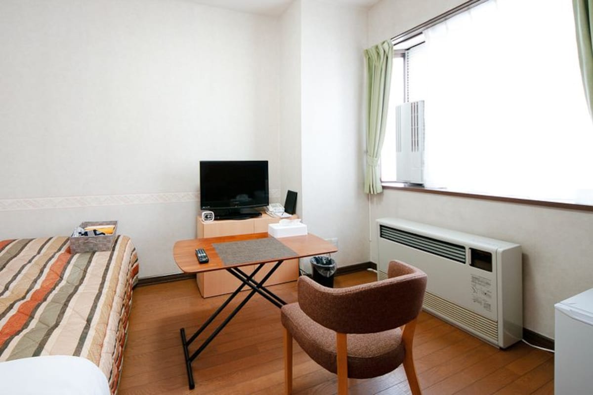 A hotel room with a bed, a desk with a television, a chair, and a radiator.