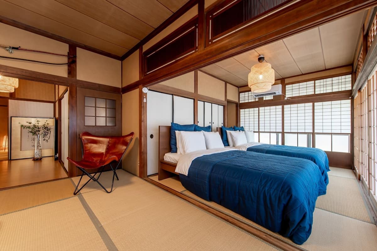 A traditional Japanese-style room with tatami flooring, a low wooden bed with blue bedding, a red leather chair, and a hanging light fixture.