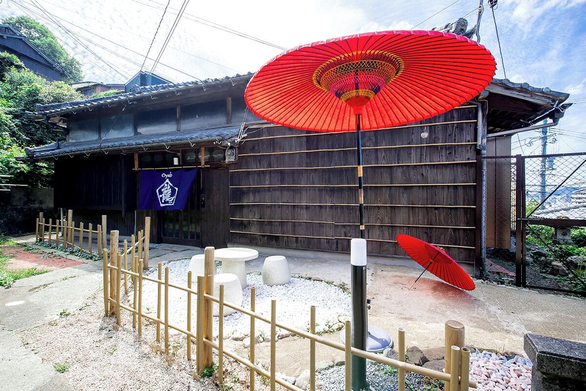 A traditional Japanese building with a wooden exterior, a red umbrella, and a small shrine-like structure with a purple banner.