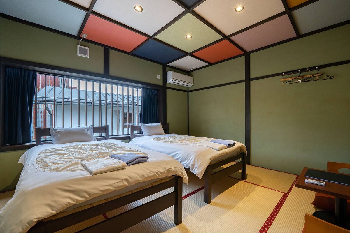 Habitación con dos camas, una ventana con rejas, un equipo de aire acondicionado y una mesita con un libro y un control remoto.