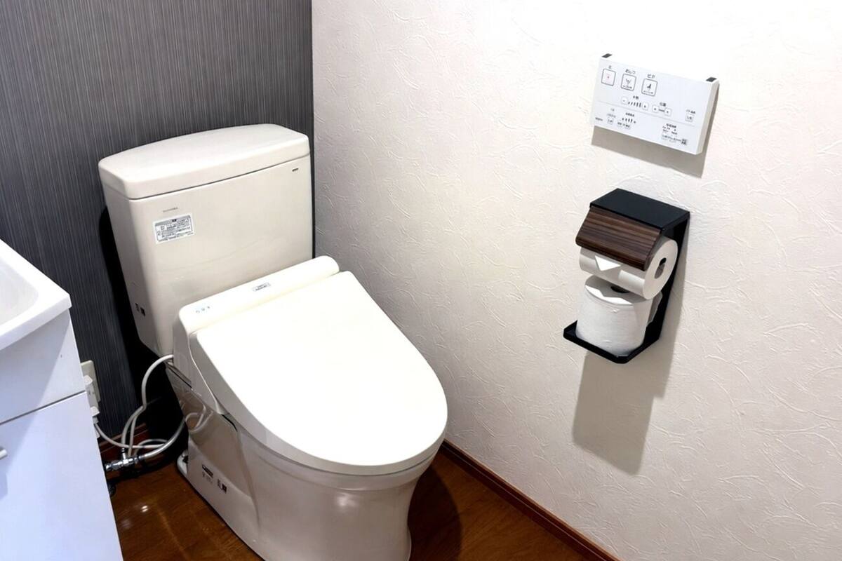 Inodoro blanco con panel de control y un dispensador de papel higiénico montado en la pared.