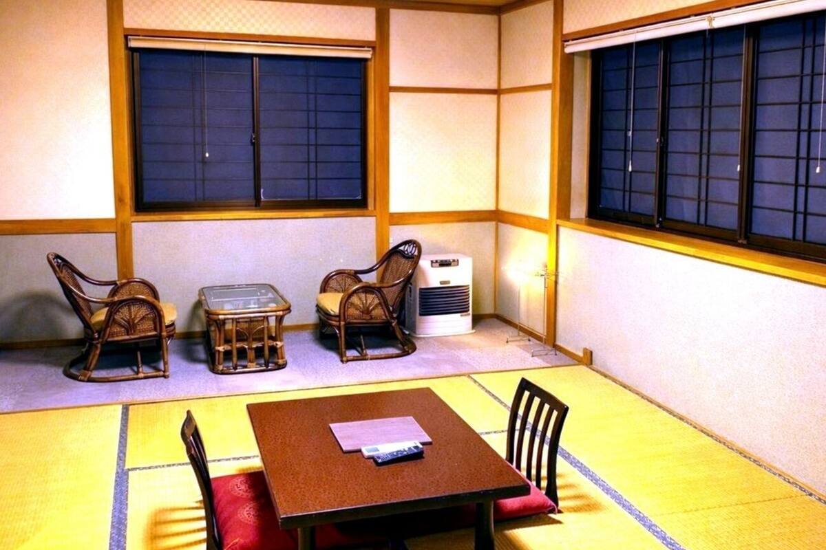 Una habitación con tatami, una mesa baja, dos sillas, un mueble pequeño y una ventana con vistas.