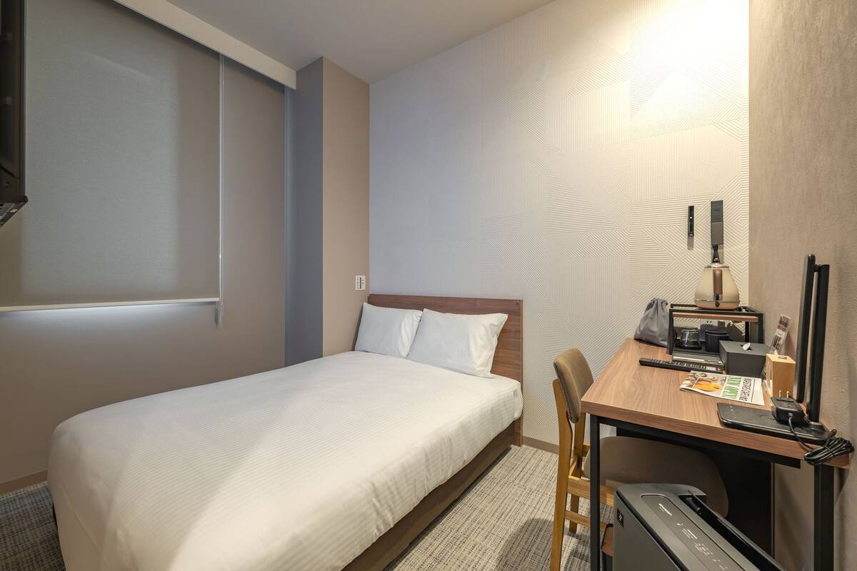 A hotel room with a bed, desk, chair, and a desk lamp.