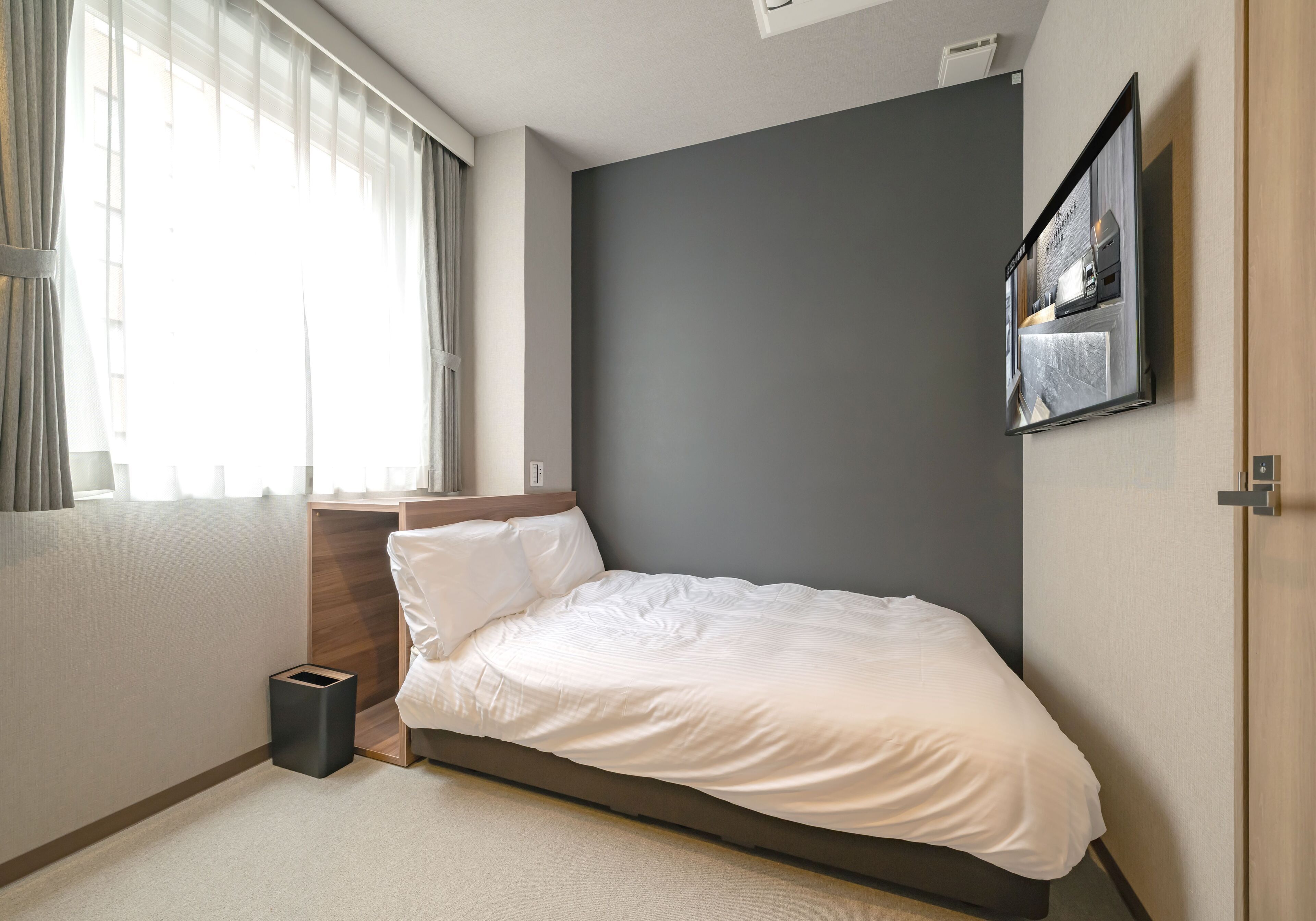 Un dormitorio con cama, mesita de noche, televisor en la pared y ventana con cortinas.