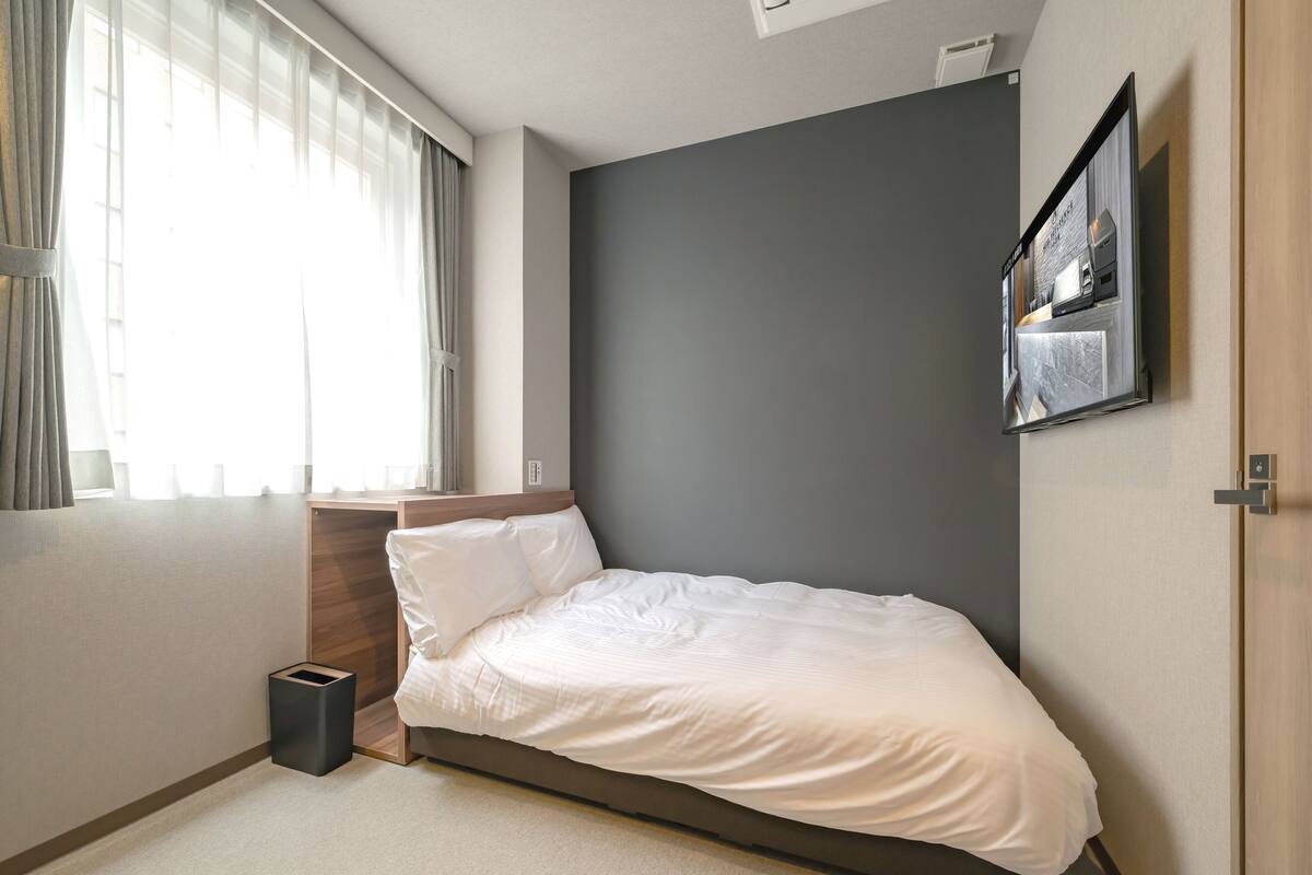 A bedroom with a bed, a bedside table, a wall-mounted TV, and a window with curtains.