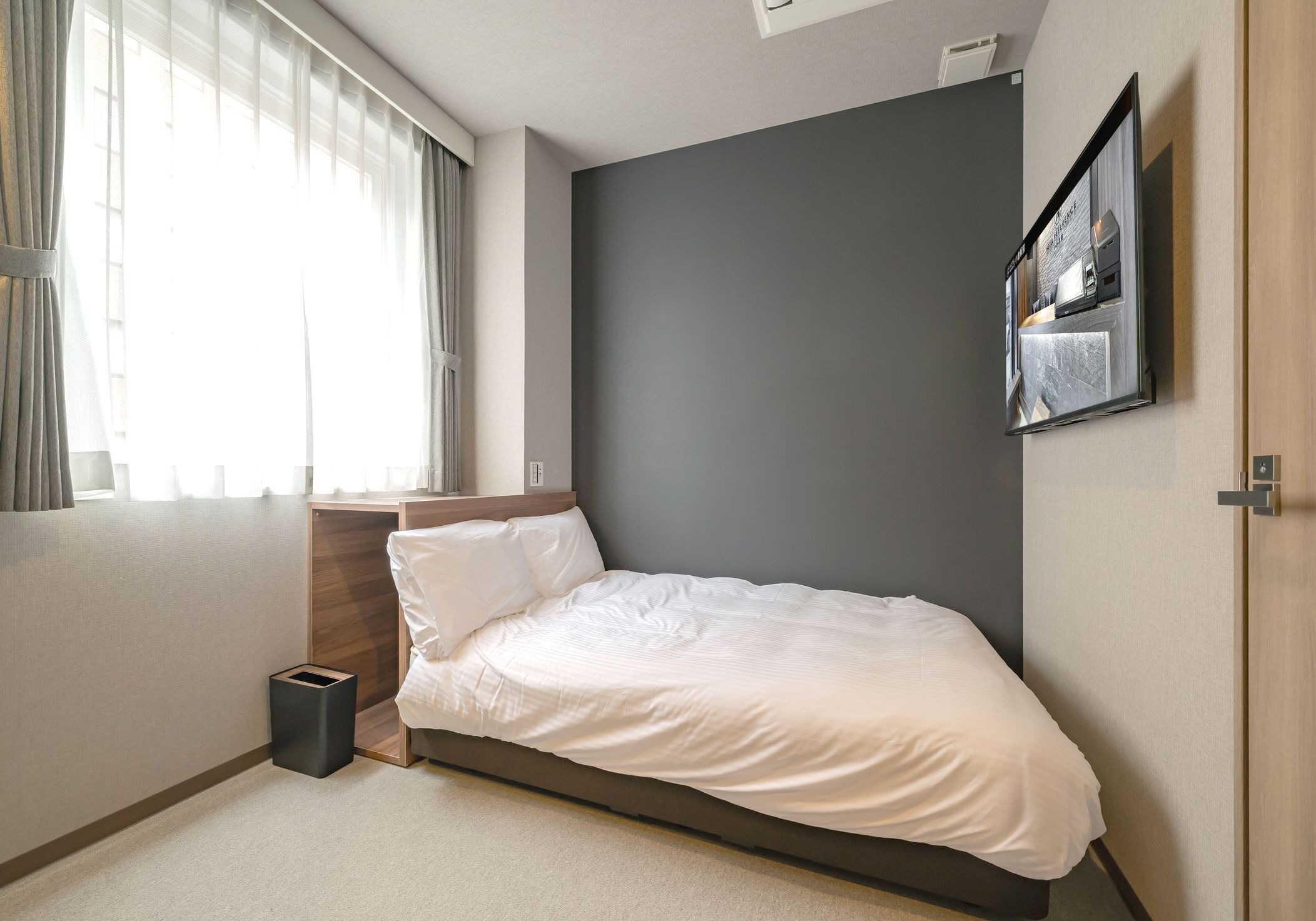 Un dormitorio con una cama, una mesita de noche, un cubo de basura, una ventana con cortinas y un cuadro colgado en la pared.