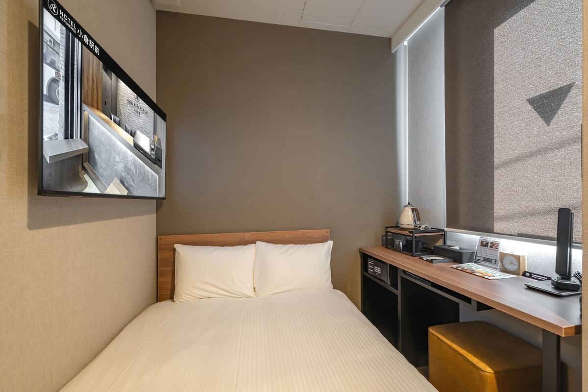 A hotel room with a bed, a desk with a computer, a window, and a wall-mounted TV.