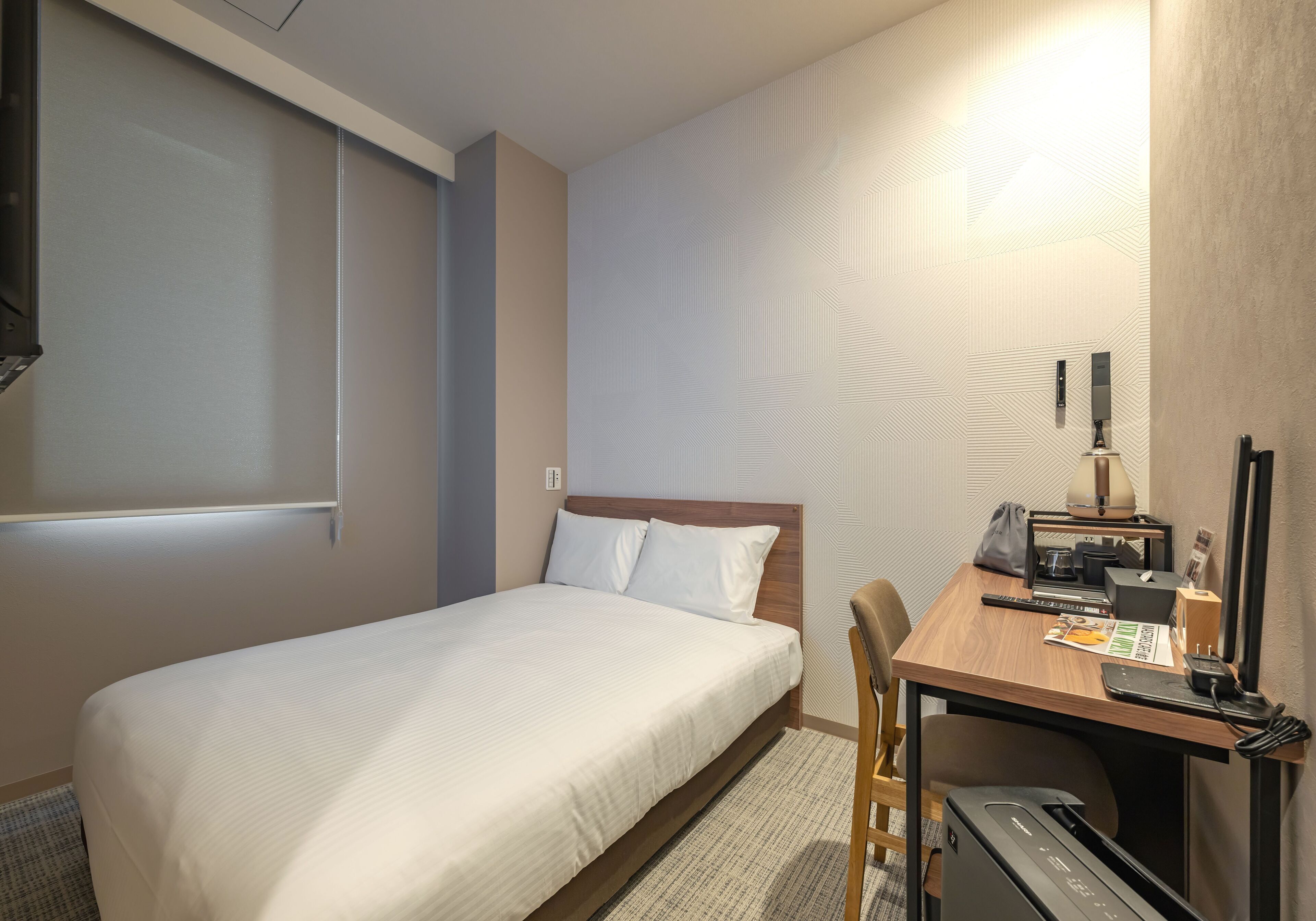 Habitación de hotel con cama, escritorio con computadora, silla, lámpara y mesita con teléfono y revista.