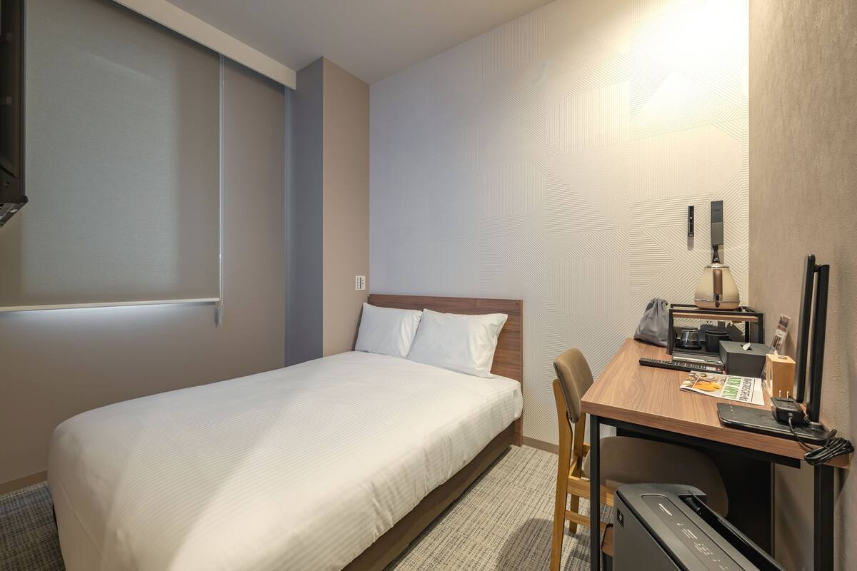 A hotel room with a bed, a desk with a computer, a chair, a lamp, and a small table with a phone and a magazine.