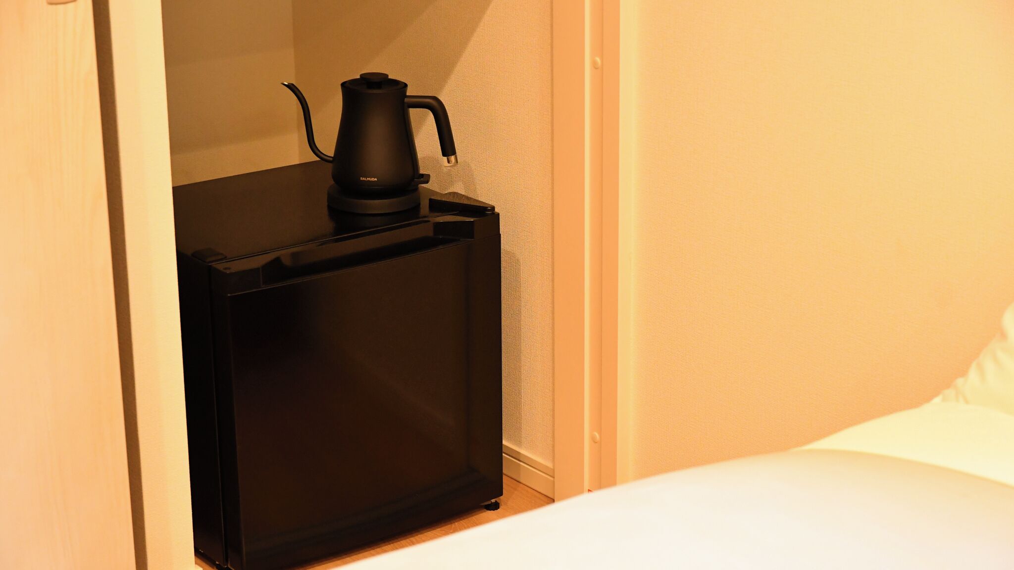A compact, dark-colored minibar with a coffee maker, located in a corner of a room with a white bed and a plain wall.