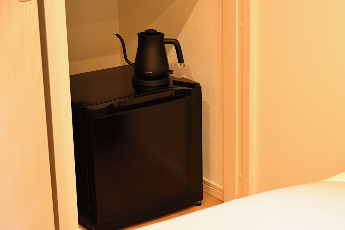 A compact, dark-colored minibar with a coffee maker, located in a corner of a room with a white bed and a plain wall.