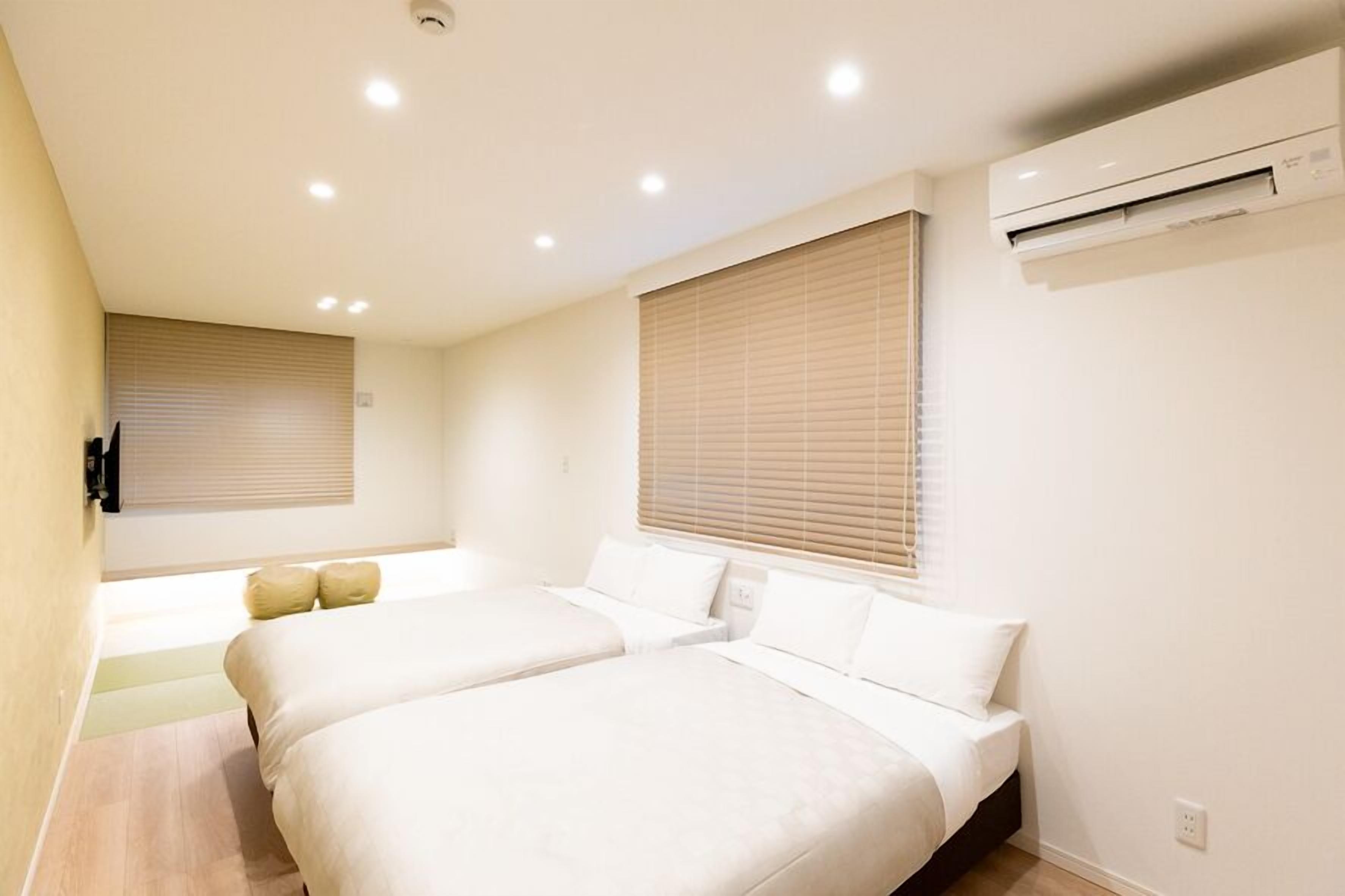 A hotel room with two beds, a wall-mounted air conditioner, and a window with blinds.