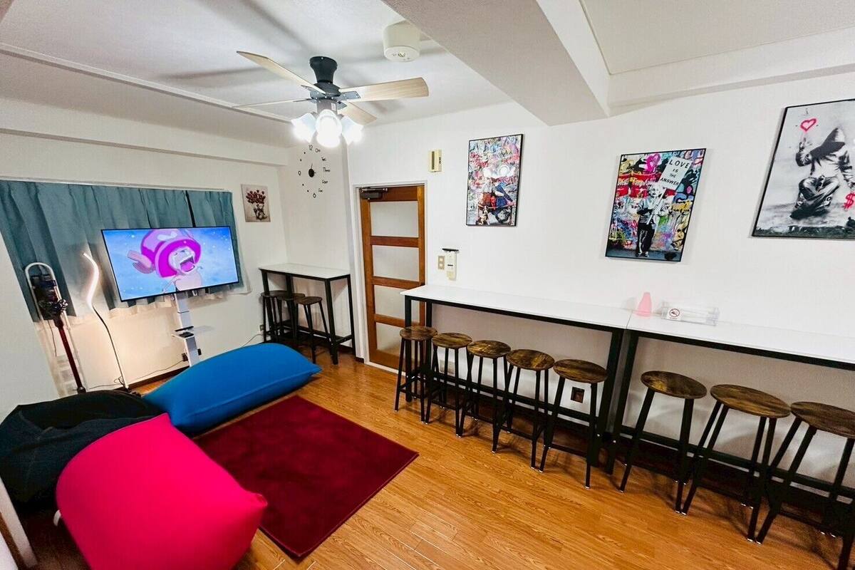A room with a TV, a ceiling fan, a bar area with stools, and artwork on the walls.