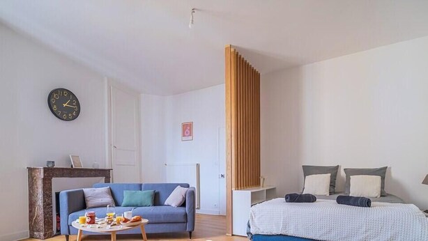 A room with a bed, a sofa, a small table with items, a clock, and a wooden partition.