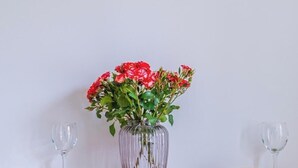 A clear glass vase with red flowers and green leaves on a table against a plain wall.