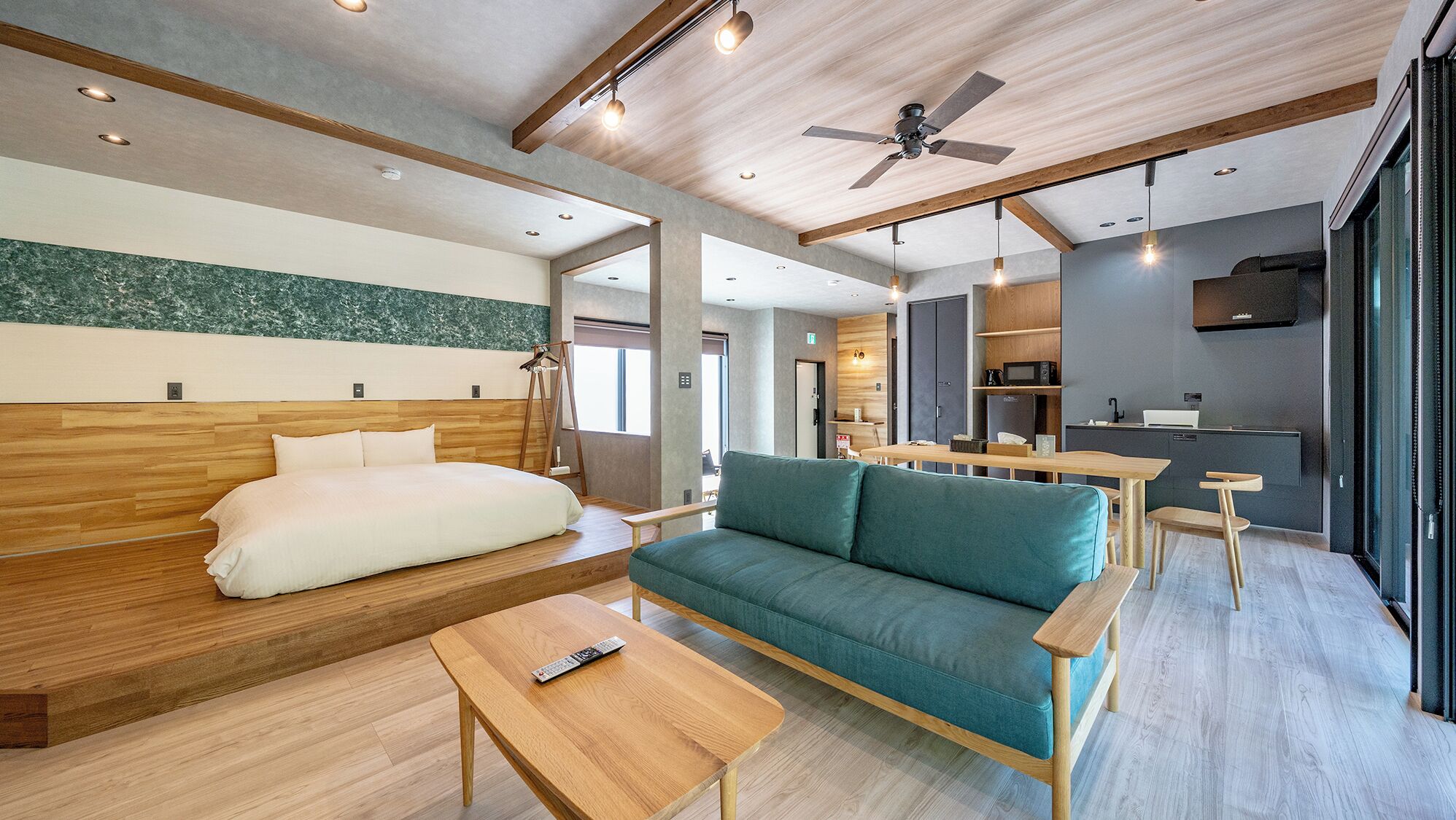 A modern hotel room with a wooden bed, a sofa, a small table, a ceiling fan, and a kitchenette.