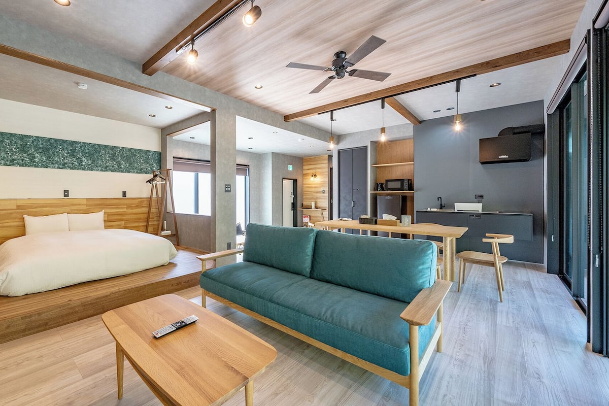 A modern hotel room with a wooden bed, a sofa, a small table, a ceiling fan, and a kitchenette.
