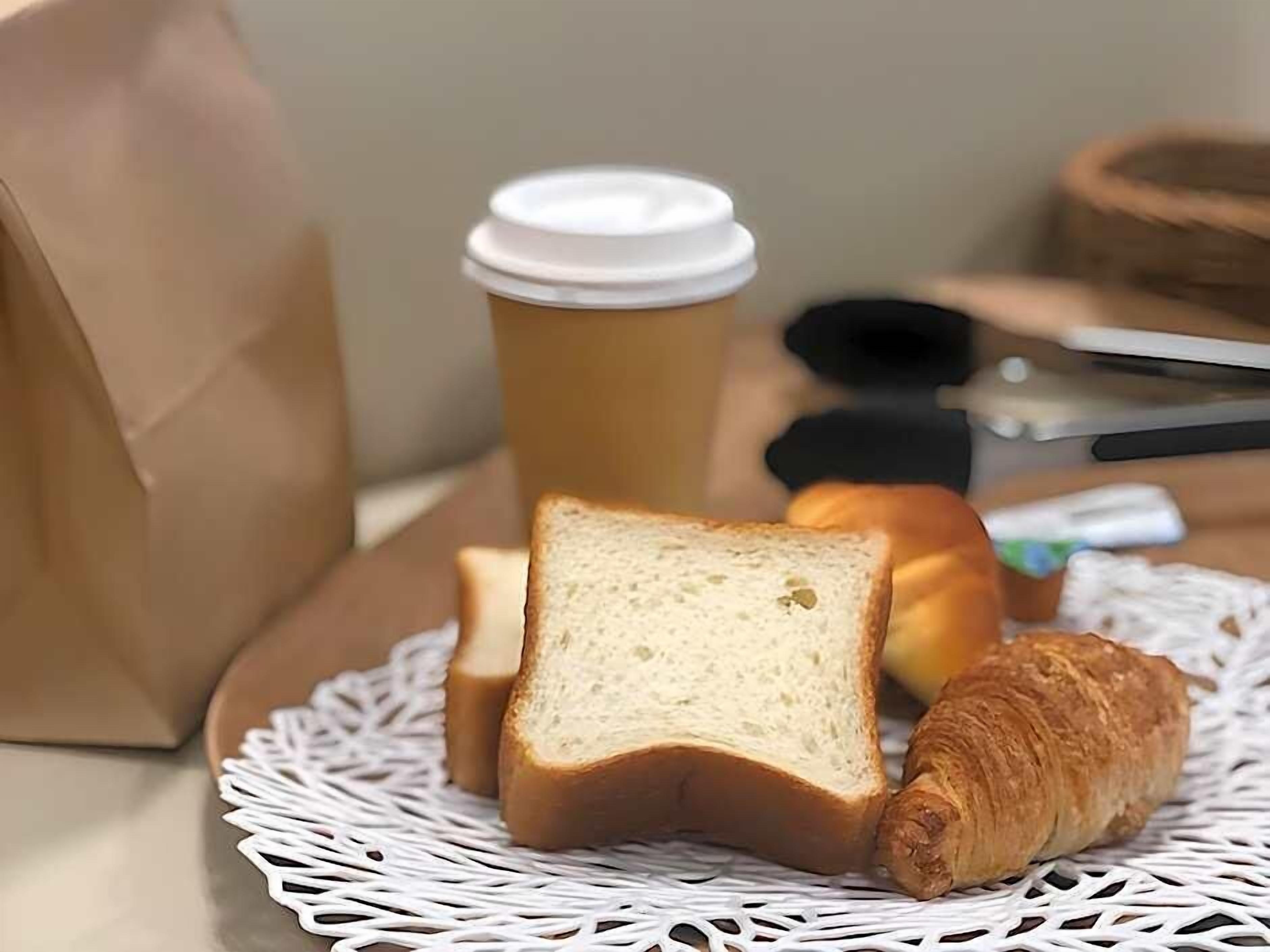 A plate with bread and a croissant on a white doily, a coffee cup, and a brown paper bag. - Business Hotel Urbanty Nishikujo (Osaka)