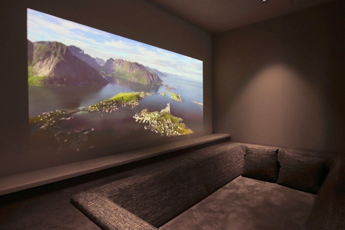 Habitación con una gran pantalla que muestra un paisaje pintoresco, un sofá de color oscuro y un estante montado en la pared.
