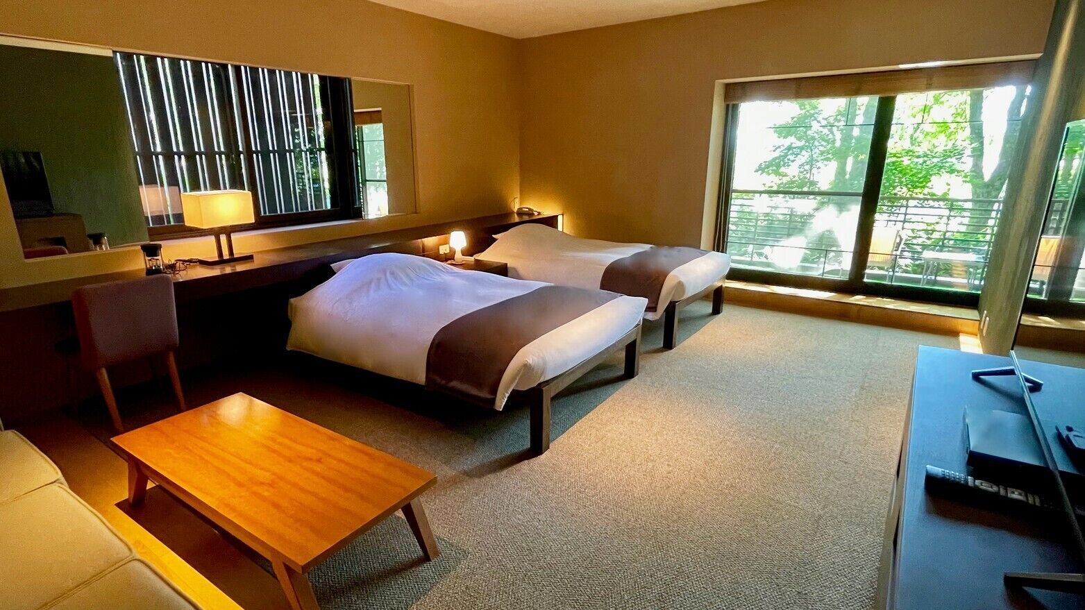 A hotel room with two beds, a desk, a chair, a lamp, and a large window with a view.