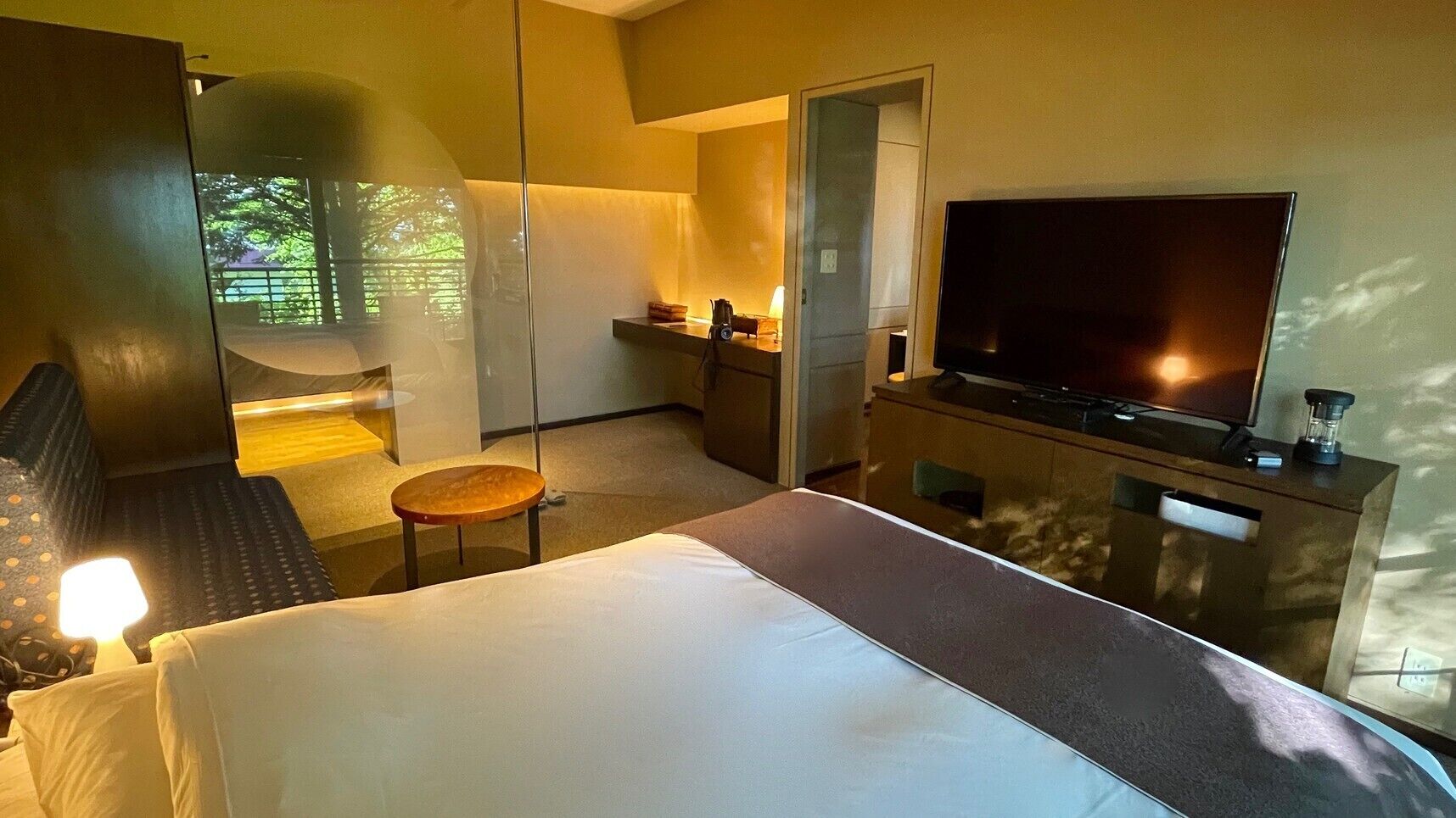 A hotel room with a bed, a TV, a desk, a chair, and a view of greenery outside.