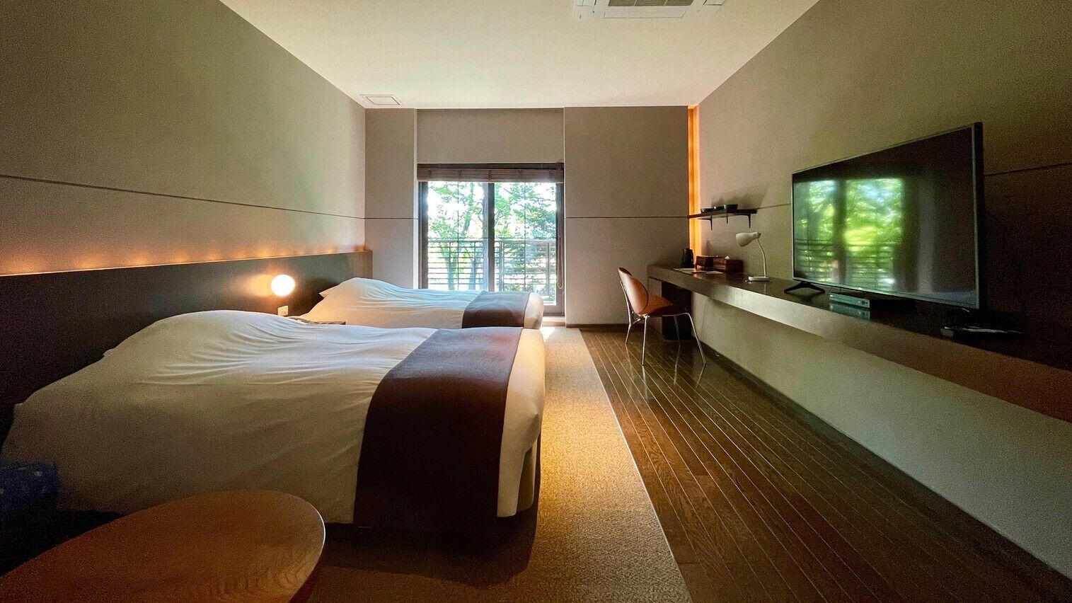 A hotel room with two beds, a desk, a TV, and a window with a view of trees.
