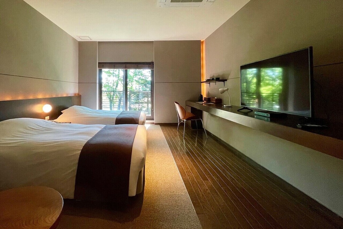 Habitación de hotel con dos camas, un escritorio, un televisor y una ventana con vistas a árboles.