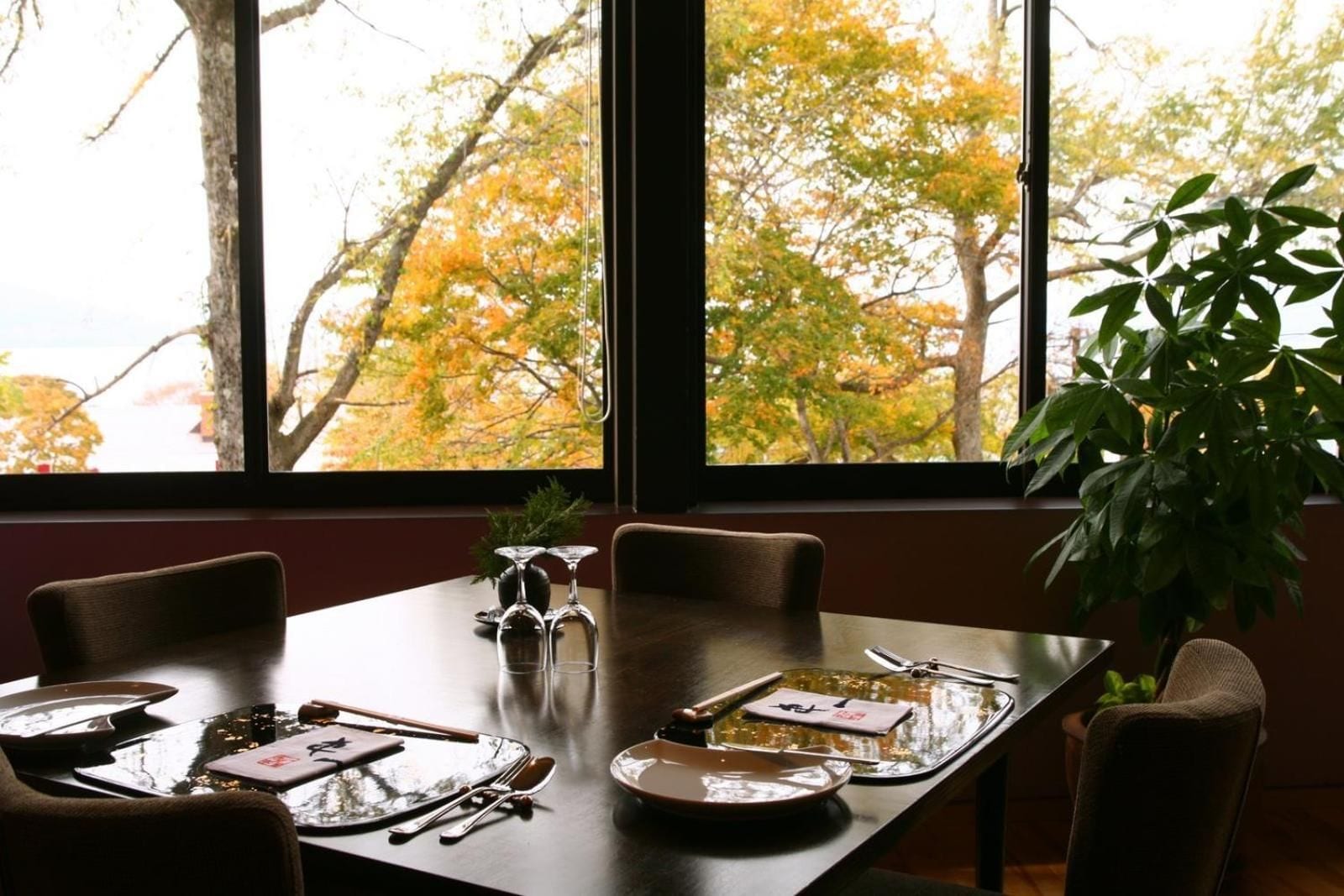 A dining table set for four with chairs, a potted plant, and a window with autumn foliage view.
