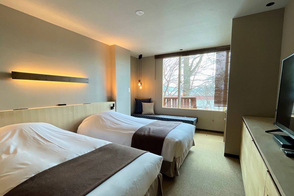 Habitación de hotel con dos camas, televisión, ventana con persianas y lámpara fijada a la pared.