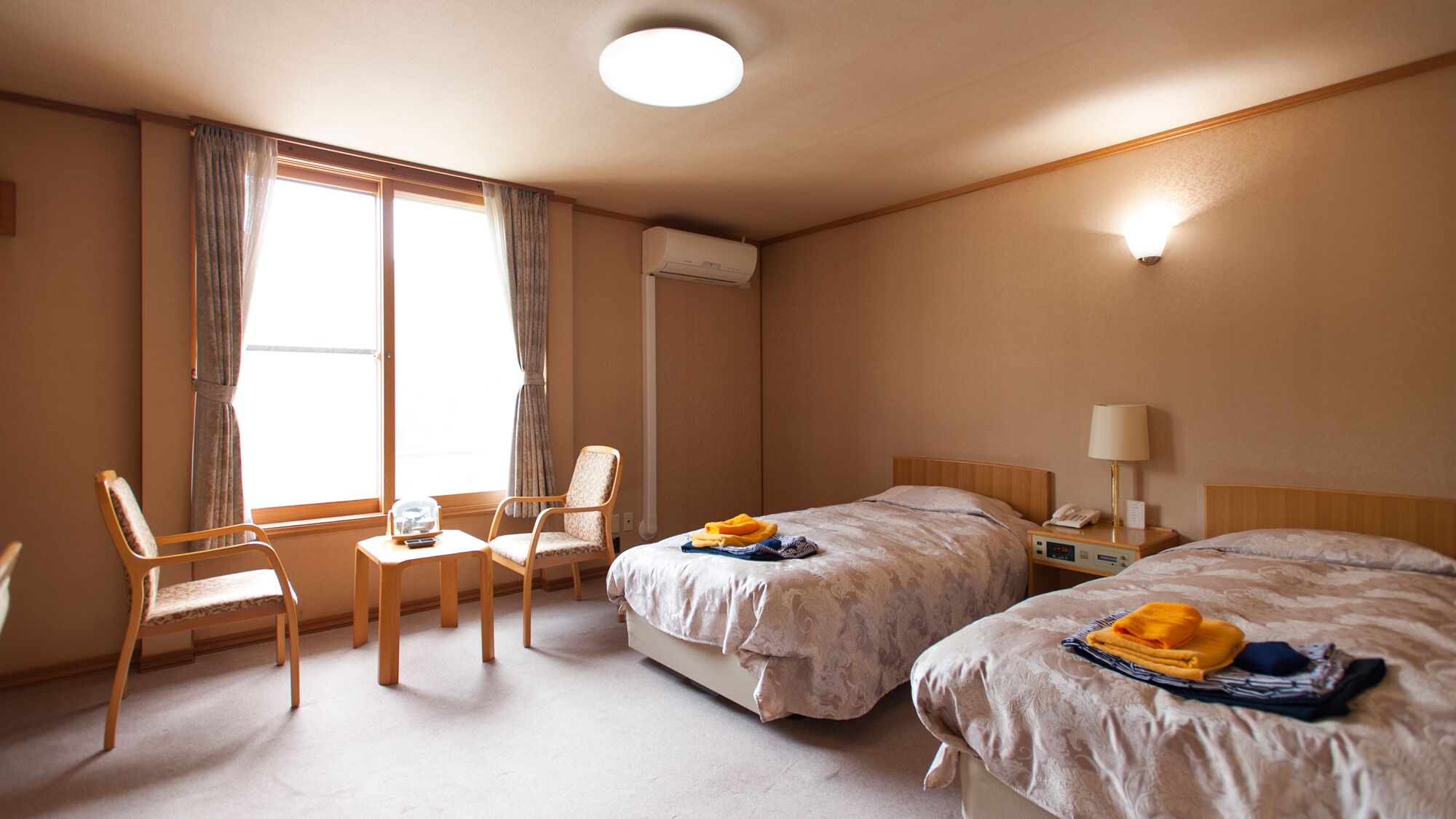 Habitación de hotel con dos camas, una silla, una mesita pequeña, un equipo de aire acondicionado y una lámpara de noche.