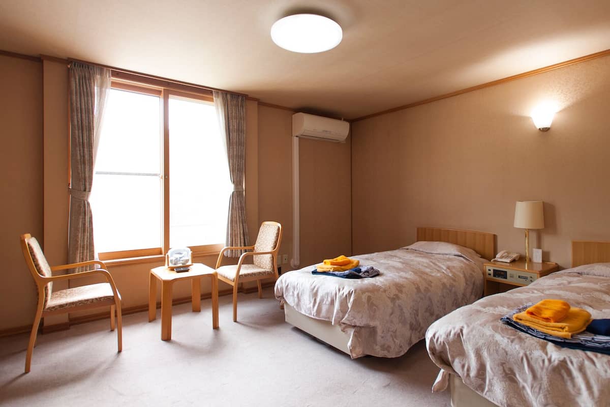 A hotel room with two beds, a chair, a small table, an air conditioning unit, and a bedside lamp.