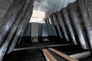 Ein schmaler, geschlossener Raum mit Metallwänden und einer geneigten Decke, der eine zentrale Metallbank und zwei Metallkästen auf dem Boden beherbergt.