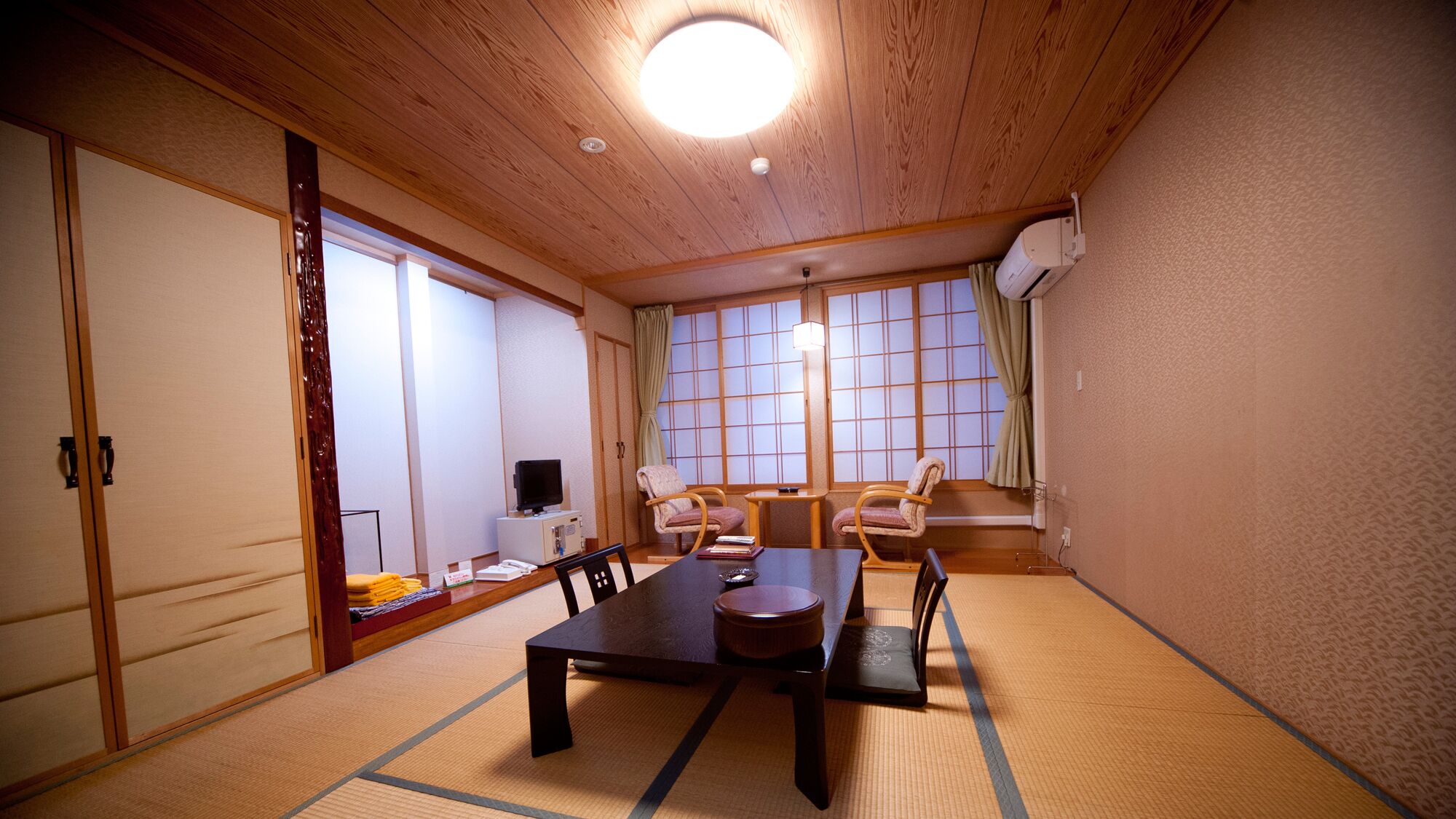 Une pièce de style japonais traditionnel, avec un sol recouvert de tatamis, une table basse, deux chaises, un téléviseur et des portes coulissantes.