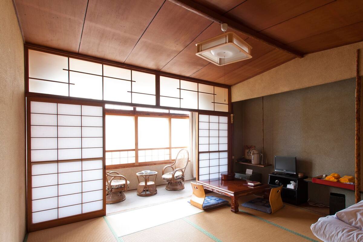 A traditional Japanese room with tatami flooring, sliding shoji doors, a low wooden ceiling, a tatami mat table, a television, and a futon bed.