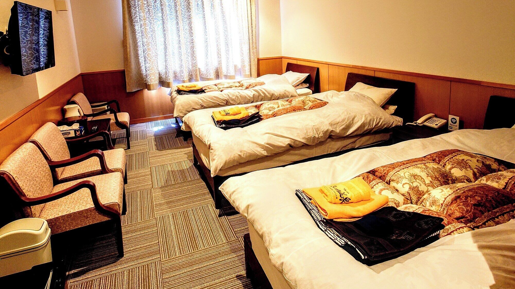 Una habitación de hotel con tres camas, un escritorio con silla, televisor y ventana con cortinas.