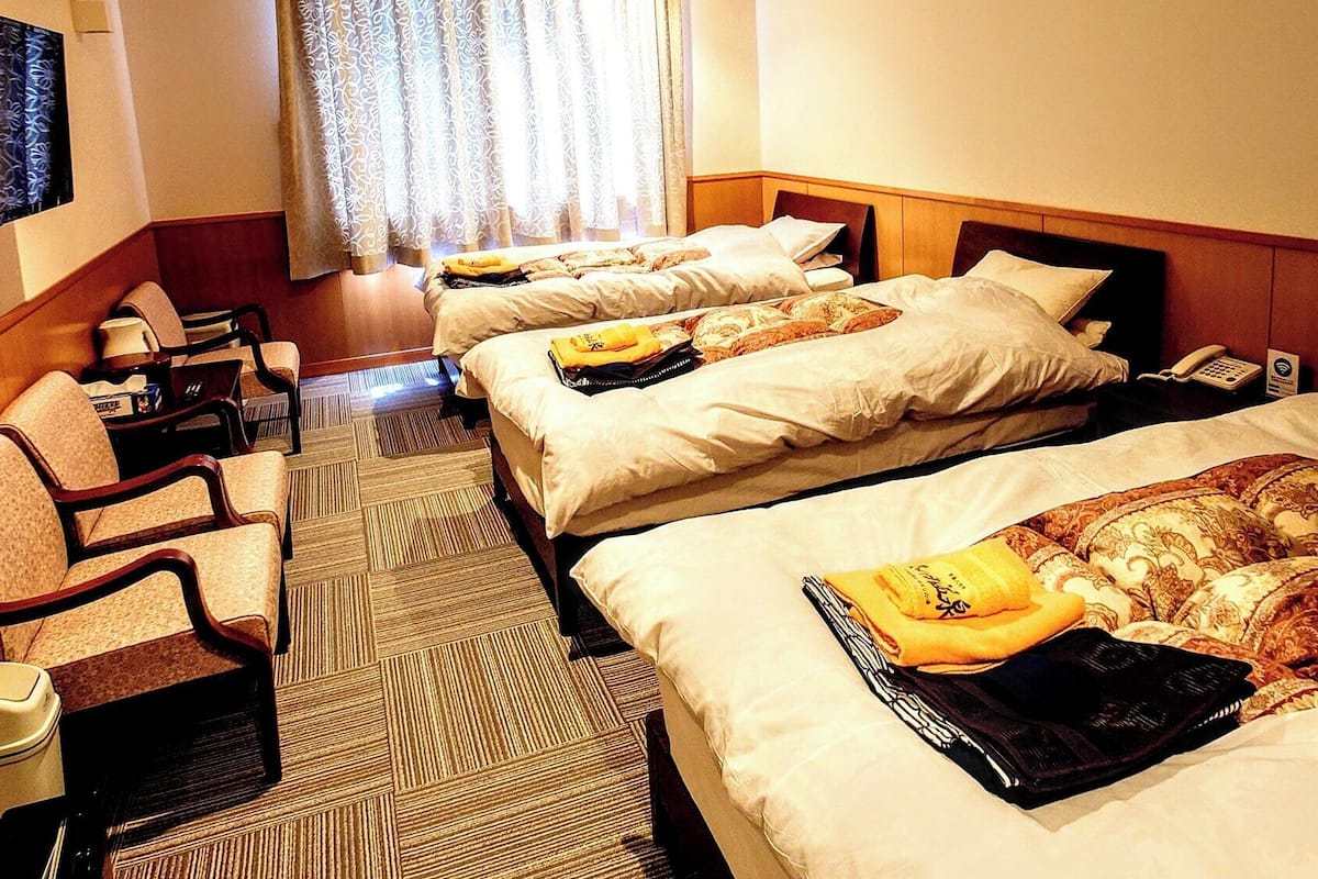 A hotel room with three beds, a desk with a chair, a TV, and a window with curtains.
