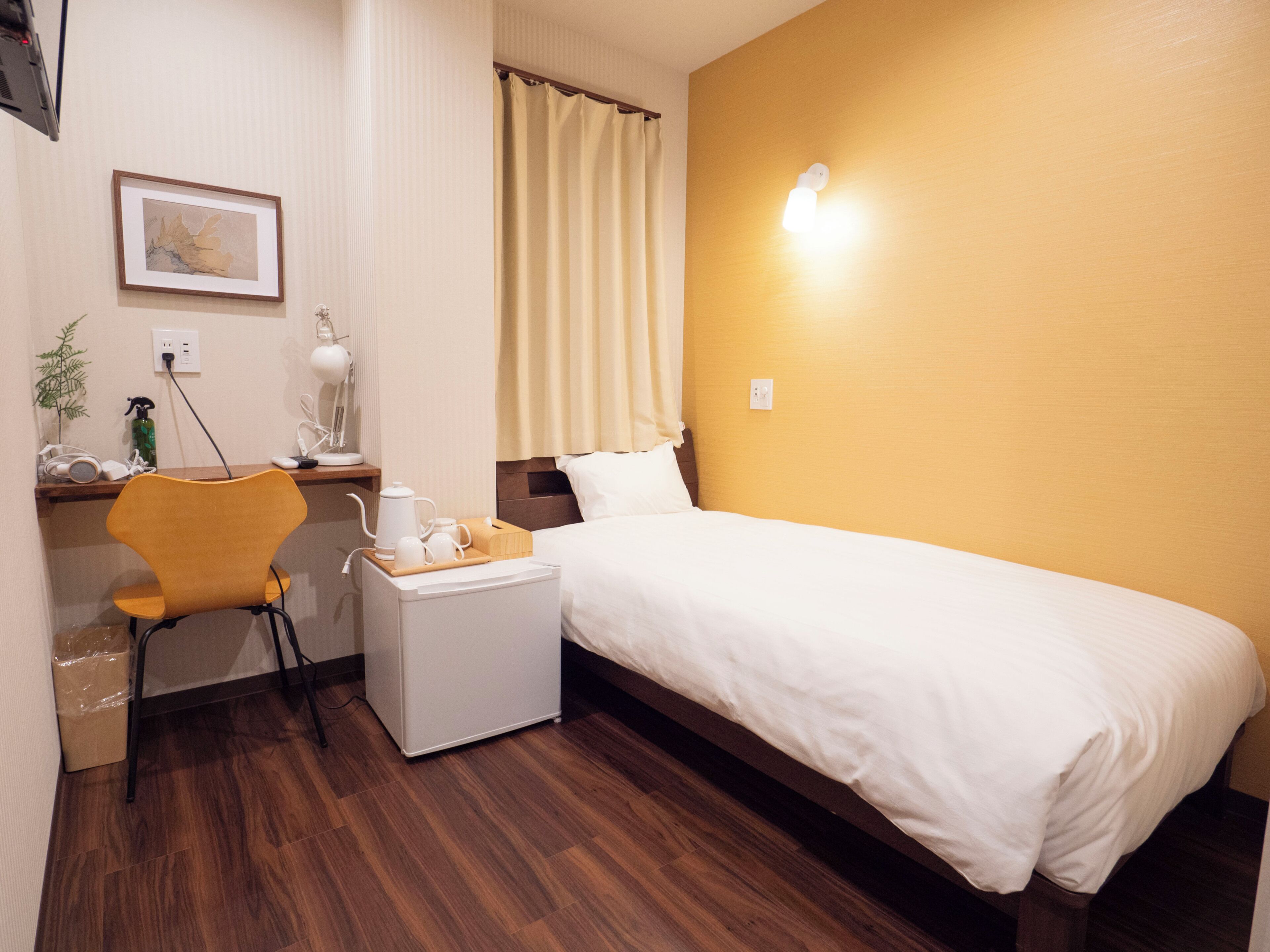 Quarto de hotel com cama, secretária com cadeira, mesinha com chaleira e chávenas, quadro na parede e janela com cortinas.