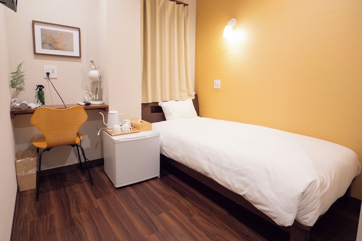 A hotel room with a bed, a desk with a chair, a small table with a kettle and cups, a picture on the wall, and a window with curtains.