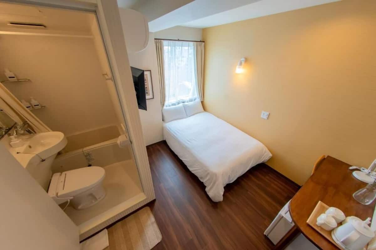 A small hotel room with a single bed, a bathroom with a shower, a sink, and a small table with a kettle and cups.