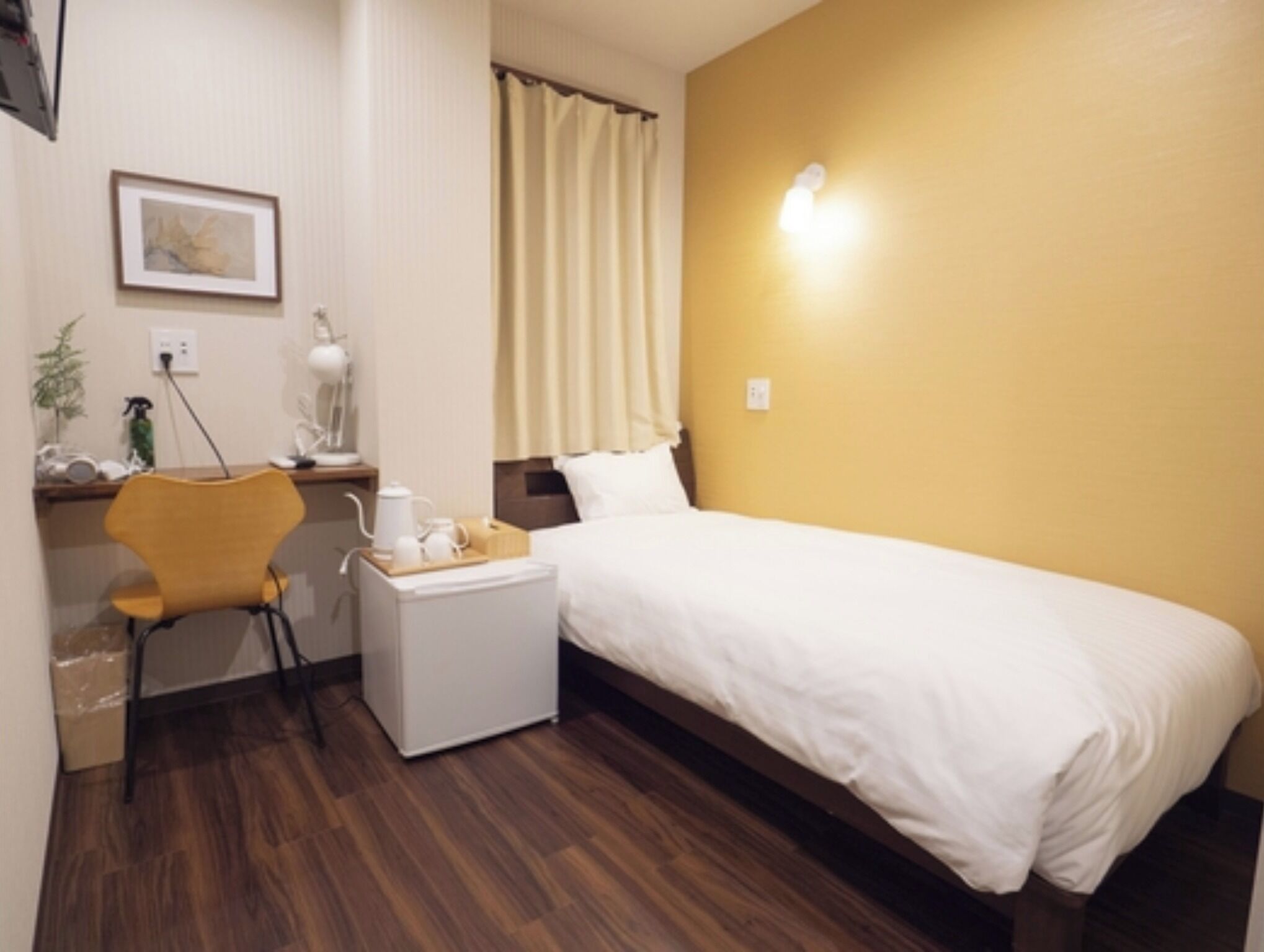 Quarto de hotel com cama, escrivaninha com cadeira, mesinhas com chaleira e chávenas, candeeiro de parede e janela com cortinas.