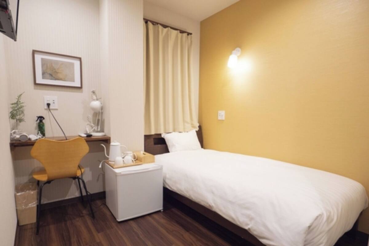 A hotel room with a bed, a desk with a chair, a small table with a kettle and cups, a wall-mounted lamp, and a window with curtains.
