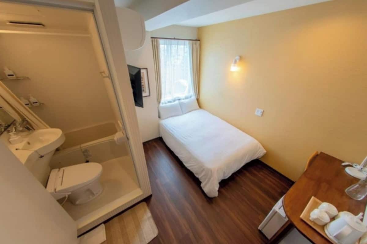A small, compact room with a single bed, a bathroom with a toilet and sink, and a small table with a lamp.