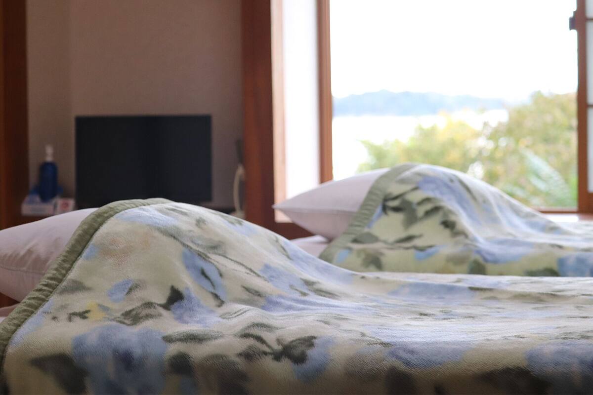 A hotel room with two beds, a TV on the wall, and a view of greenery outside.