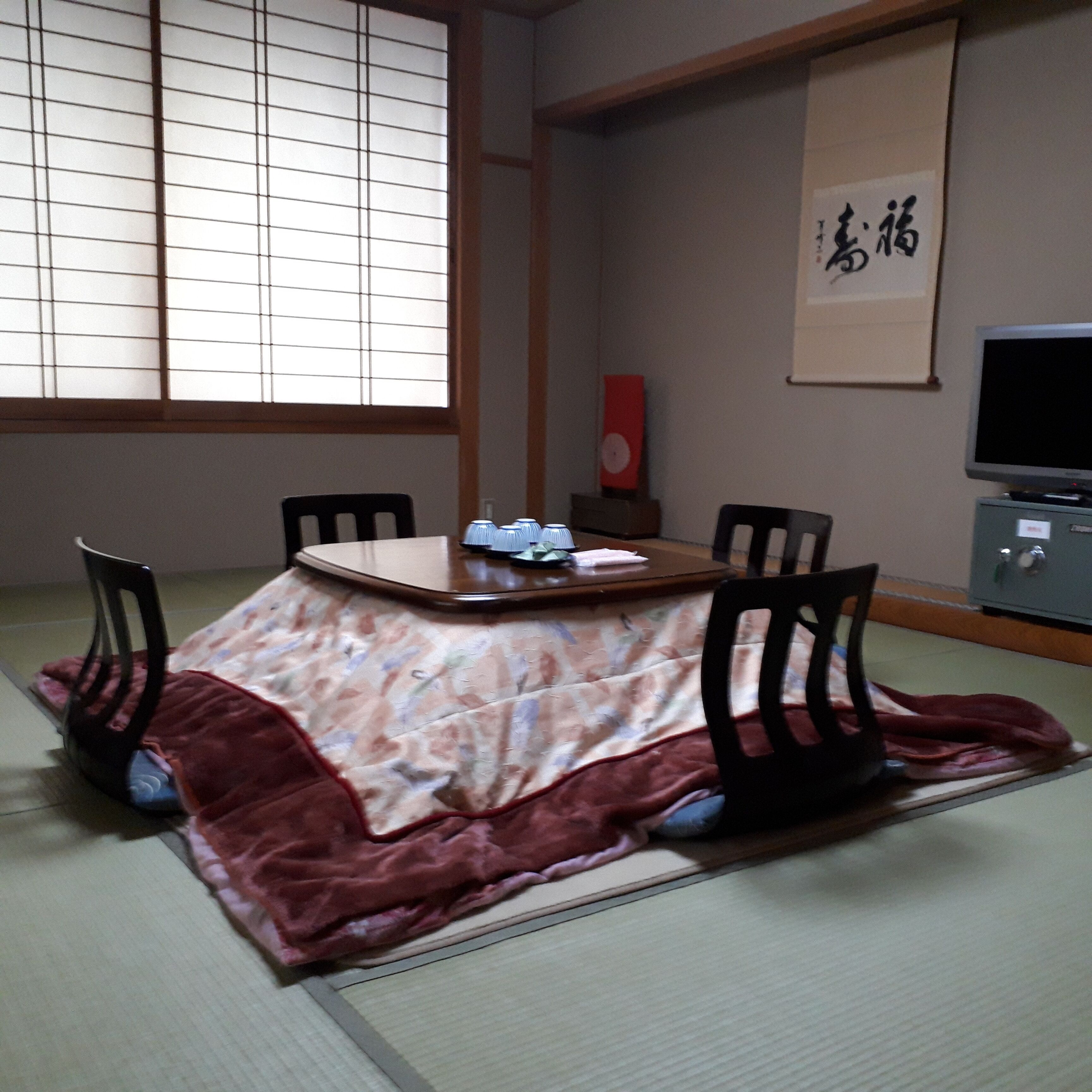 Et traditionelt japansk rum med tatamimåtter, et lavt bord med dug, fire stole, et fjernsyn og et indramet kalligrafiværk på væggen.