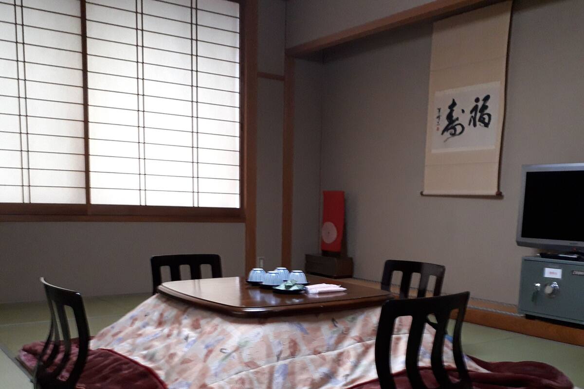 Habitació japonesa tradicional amb sòcols de tatami, una taula baixa amb estovell, quatre cadires, un televisor i una obra de cal·ligrafia emmarcada a la paret.