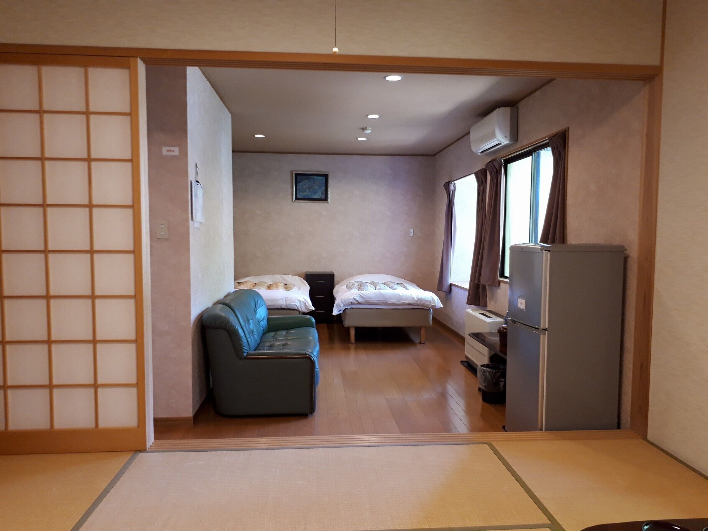Quarto compacto com cama tipo futon, um pequeno sofá, geladeira e janela com cortinas.