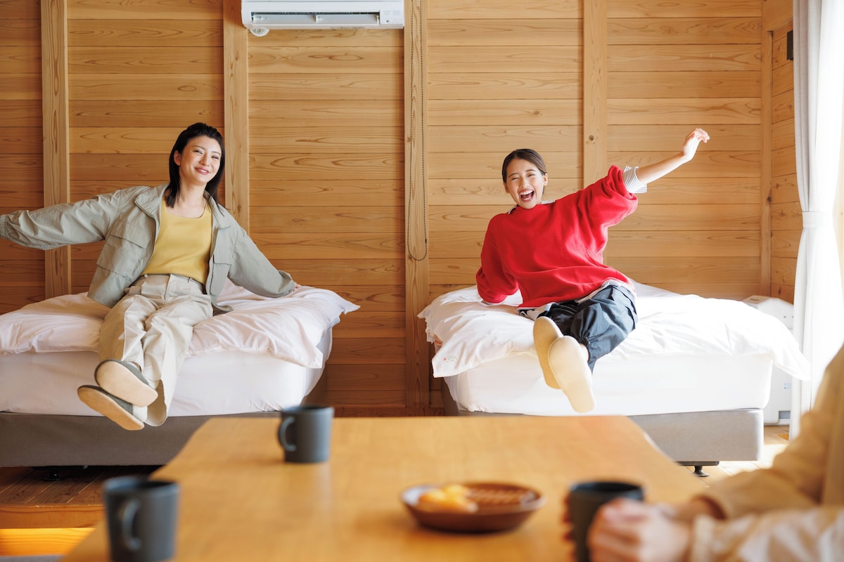 A room with wooden walls, two beds, a table with a bowl and mugs, and an air conditioning unit.