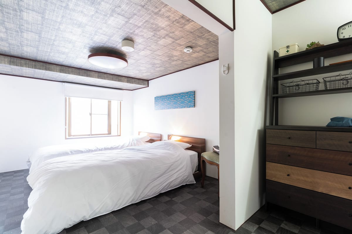 A bedroom with a large bed, a nightstand, a dresser, a ceiling light, a window, and a wall-mounted shelf with items.